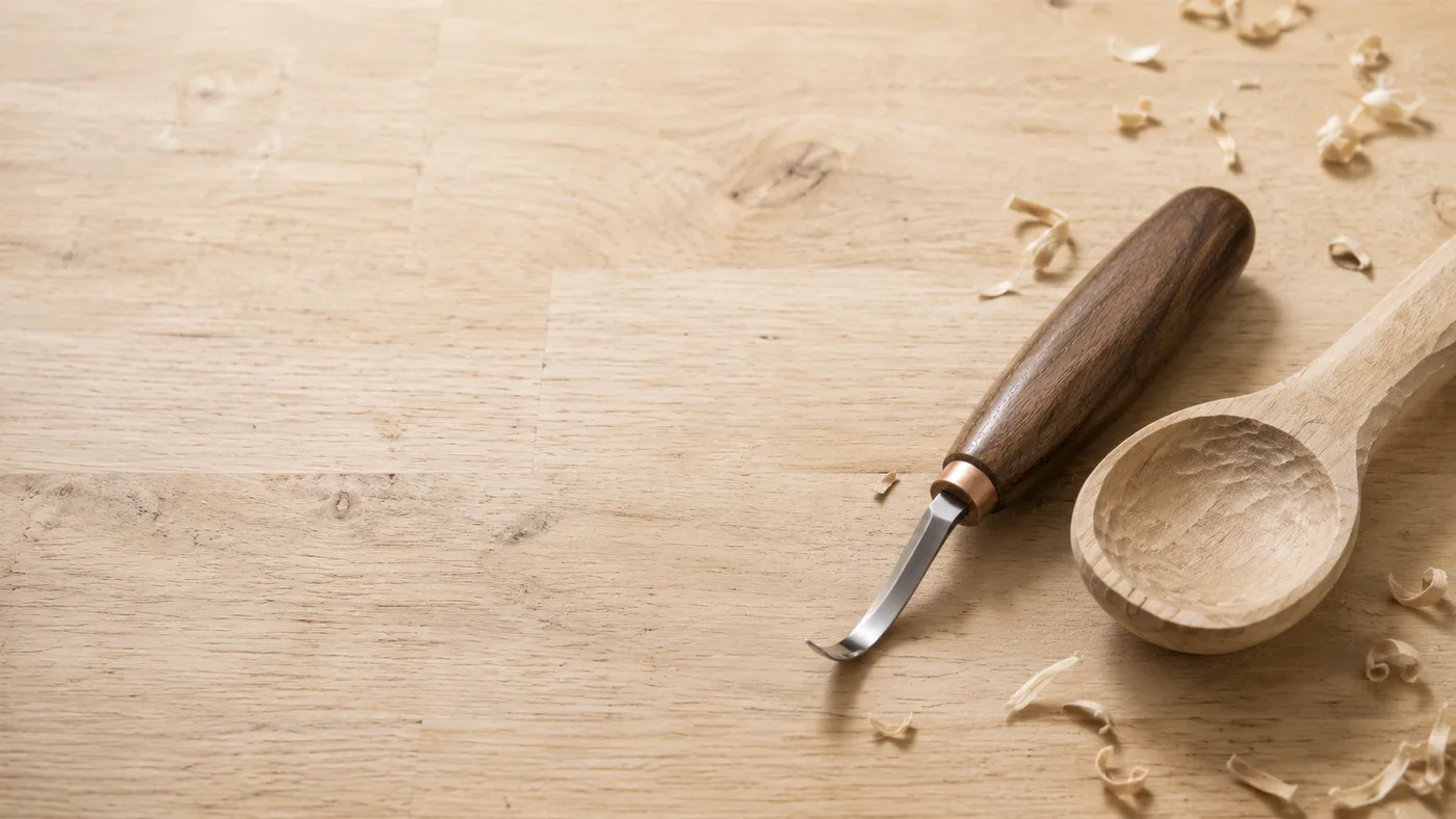 A walnut-handled hook knife resting beside a half-carved wooden spoon on a pale workbench, shavings scattered around
