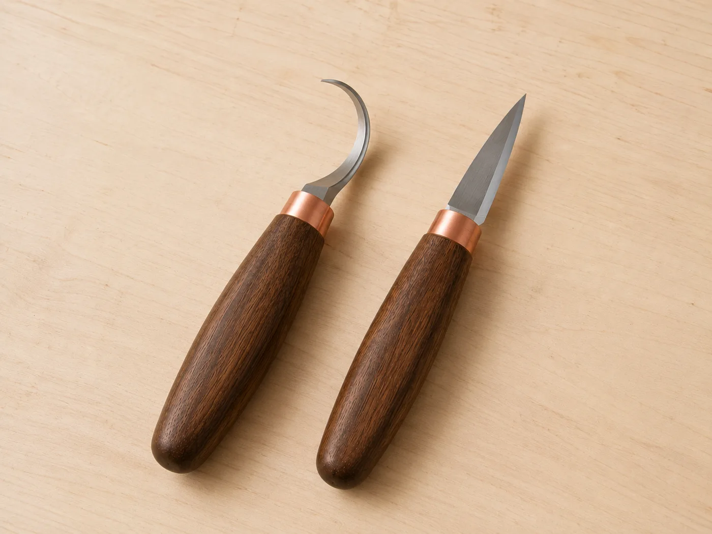 The hook knife and the straight sloyd knife placed side by side on light wood, the contrast between their blade shapes clearly visible