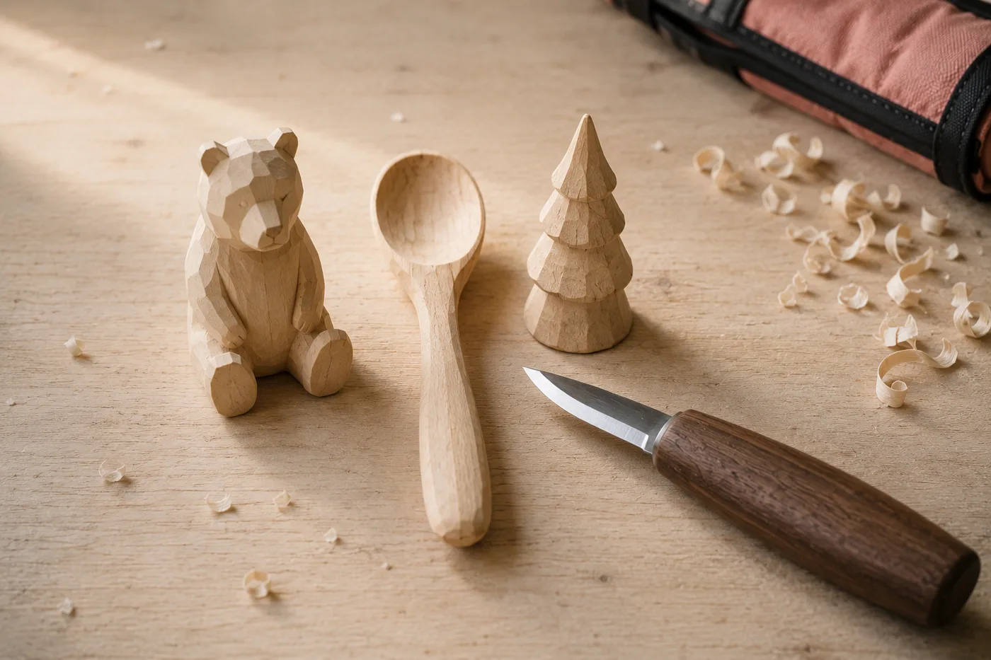 Three finished whittling projects on a pale workbench — a small bear, a spoon and a fir tree — with a carving knife beside them